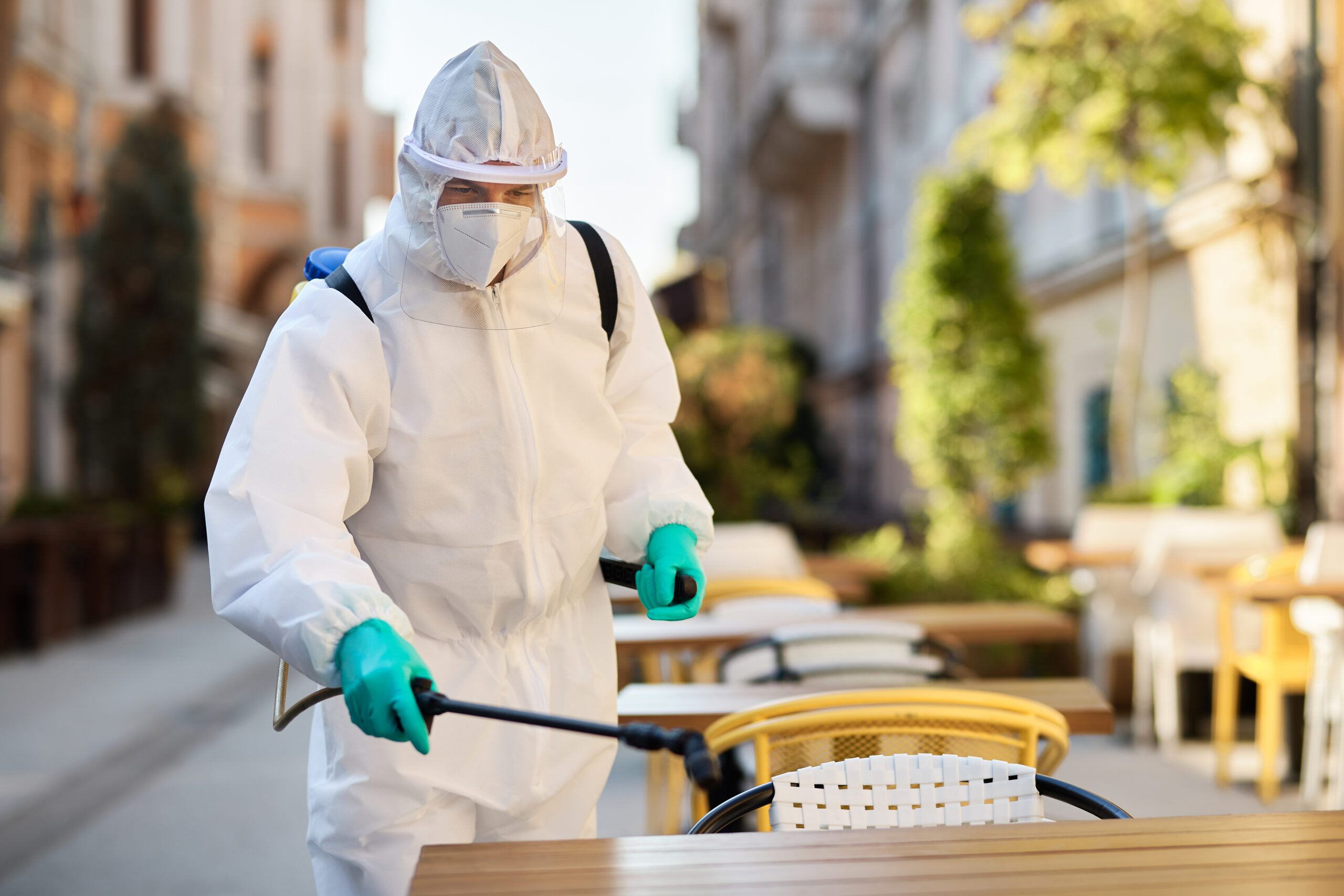 Sanitation worker in protective suit doing disinfection of an outdoor cafe due to coronavirus pandemic.