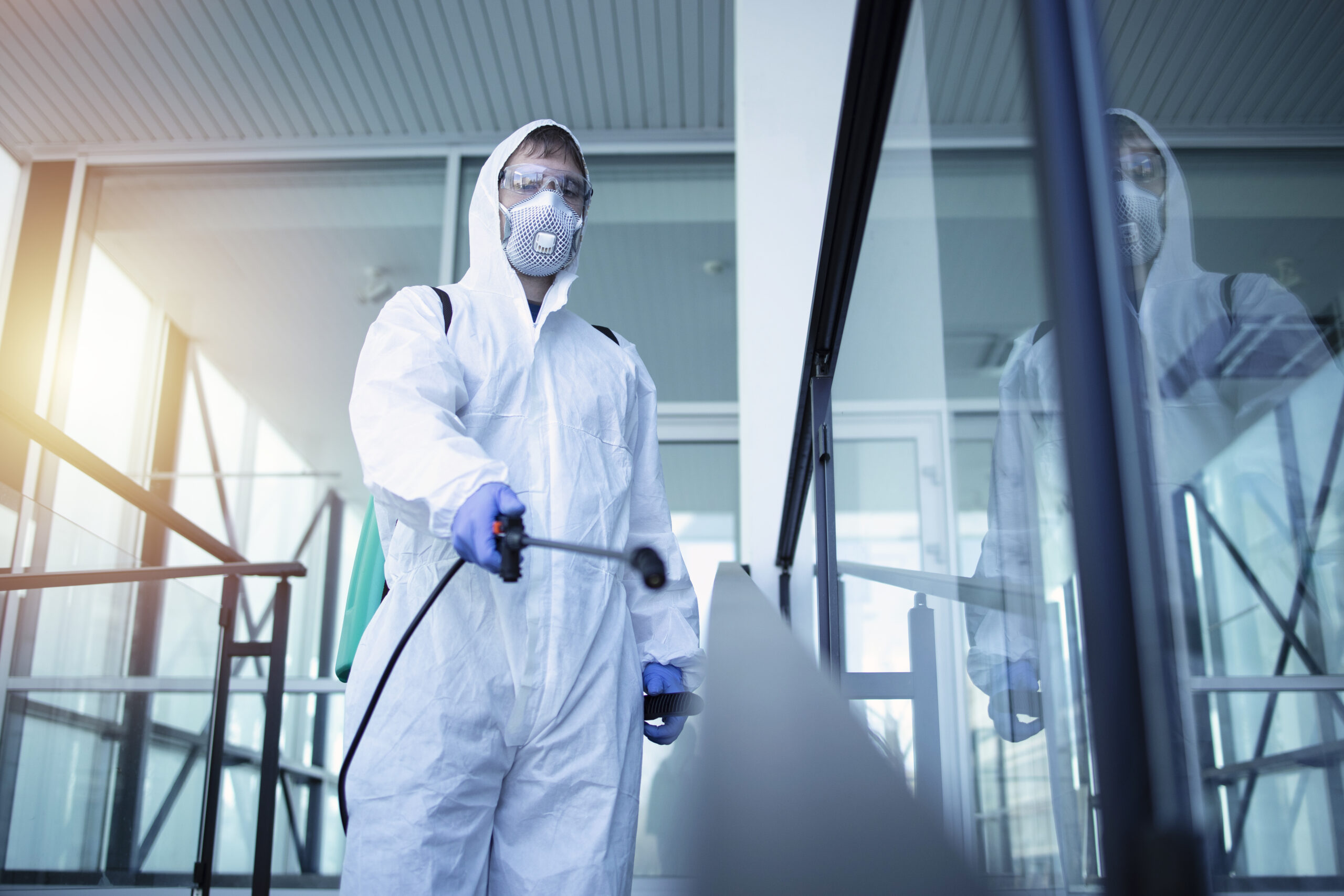 Person in white chemical protection suit doing disinfection of public areas to stop spreading highly contagious corona virus.