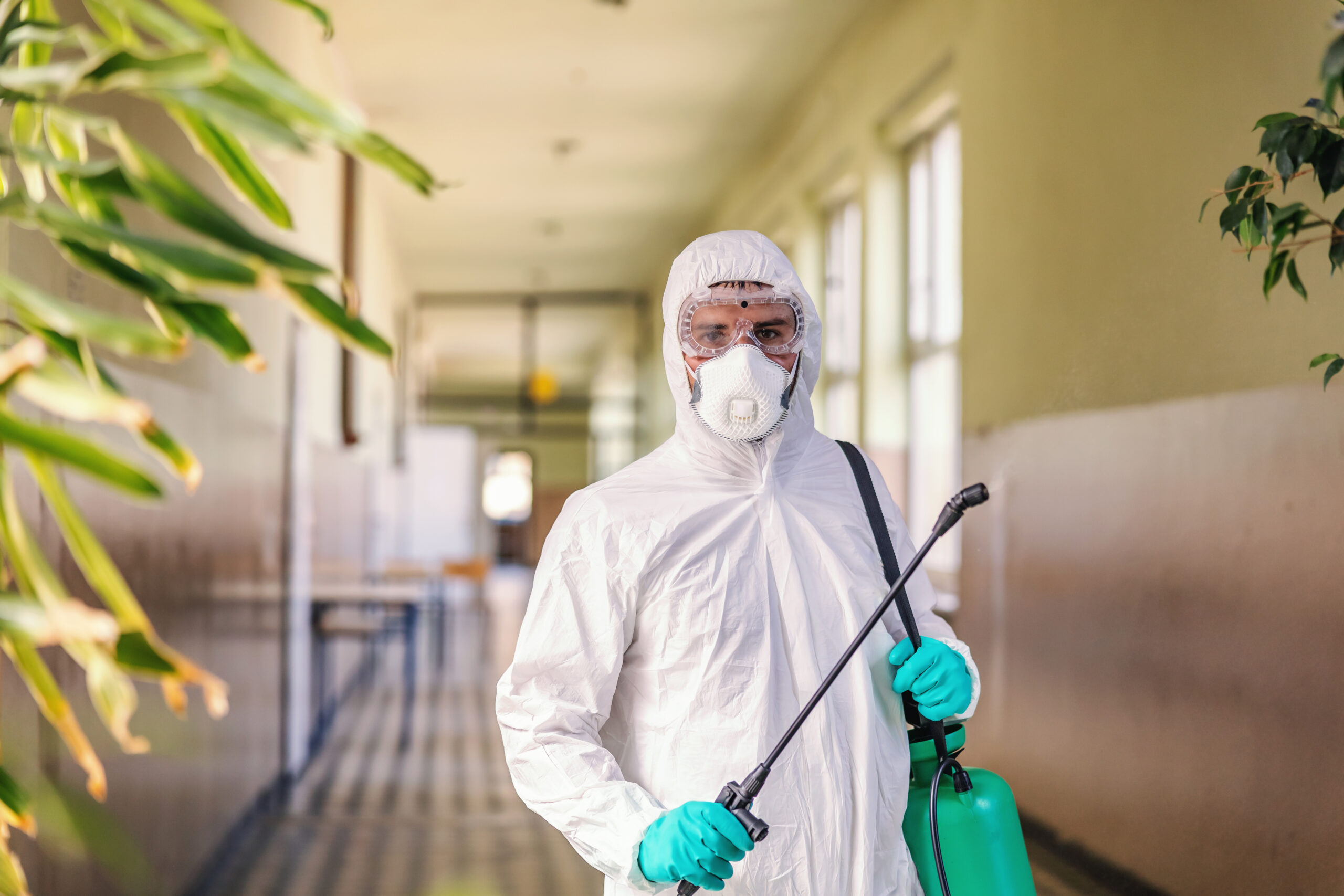 Portrait of worker in sterile white uniform, with face mask and rubber gloves on standing in hallway in school and holding sprayer with disinfectant.