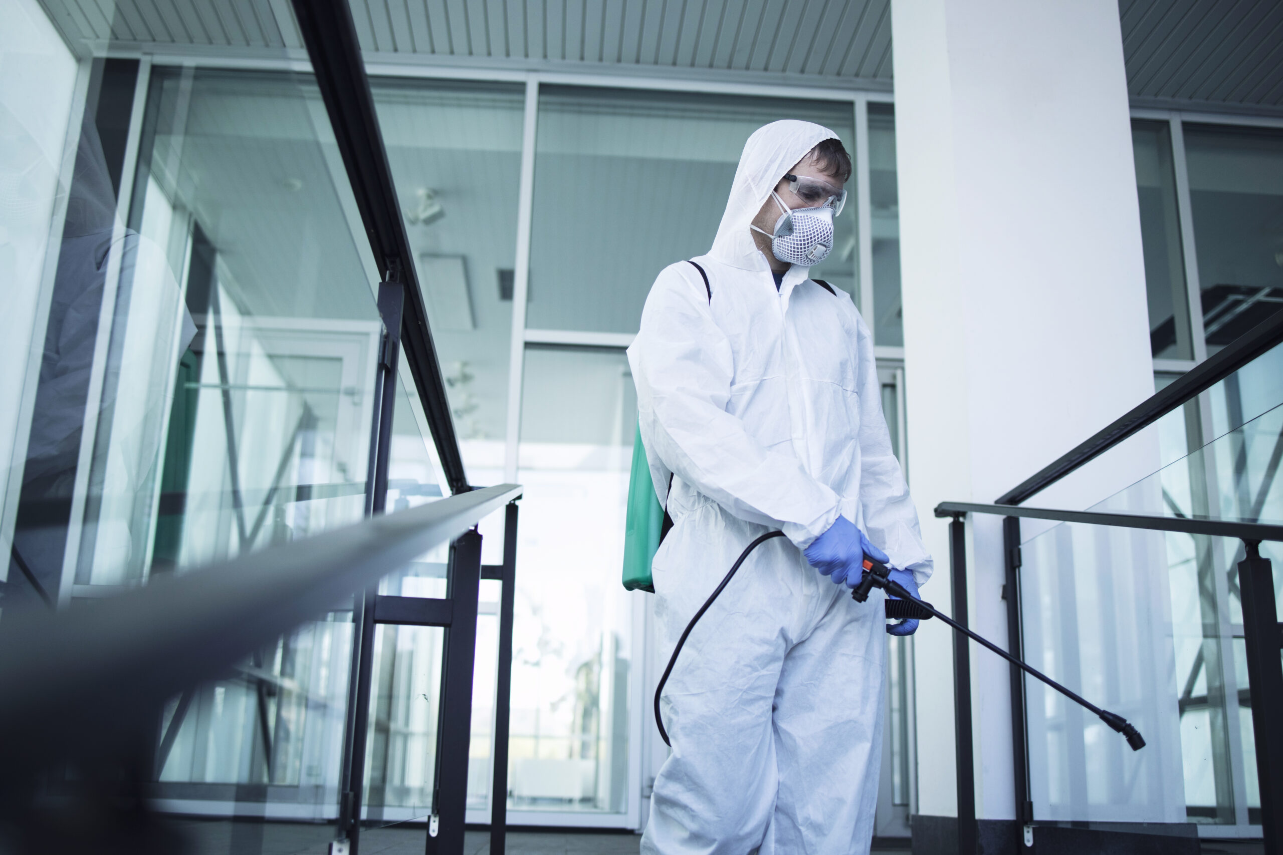 Person in white chemical protection suit doing disinfection of public areas to stop spreading highly contagious corona virus.